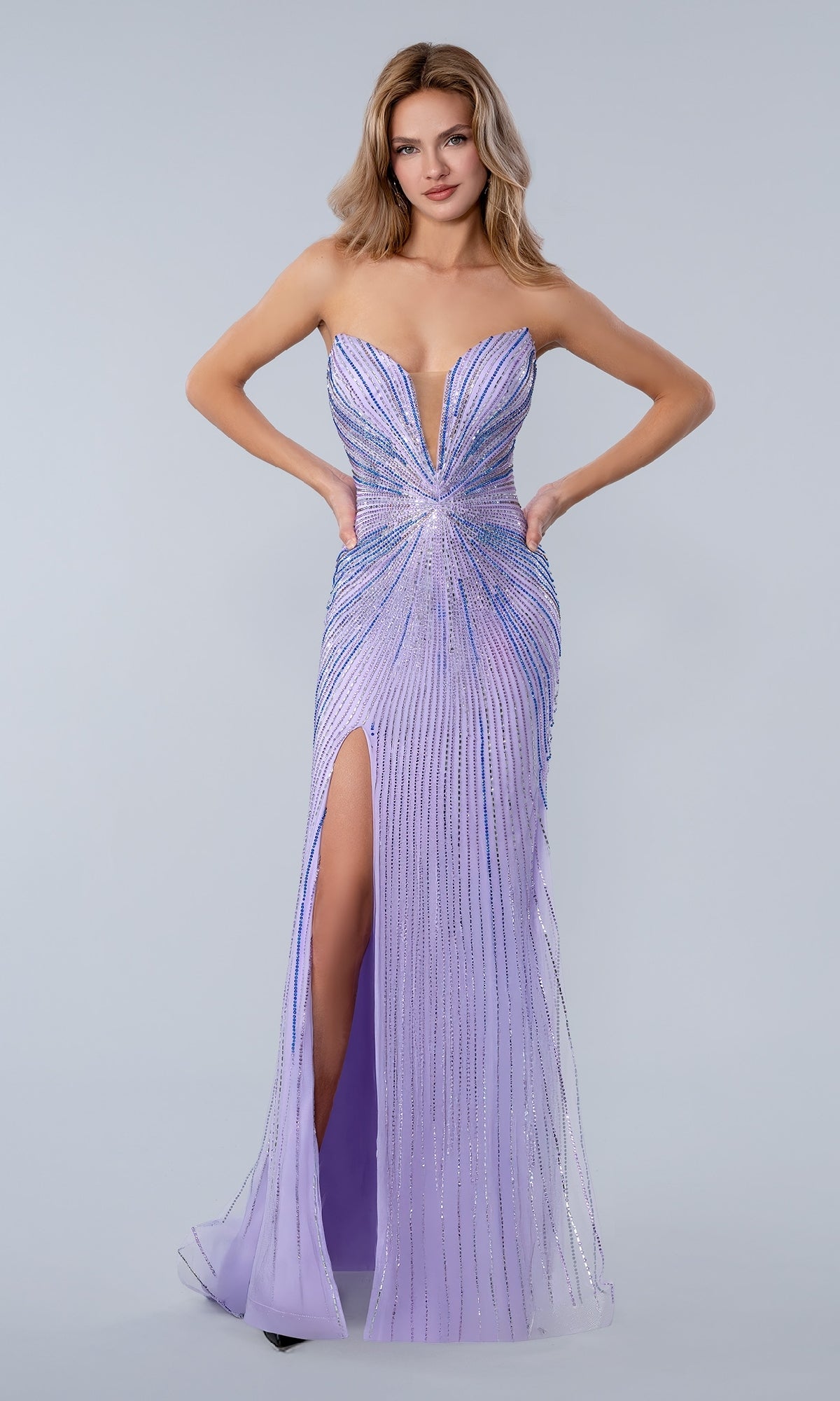 A woman models the Long Prom Dress Stella Couture 26163 in lilac, featuring a strapless design, deep neckline, thigh-high slit, and sparkling rhinestone embellishments against a light grey background.